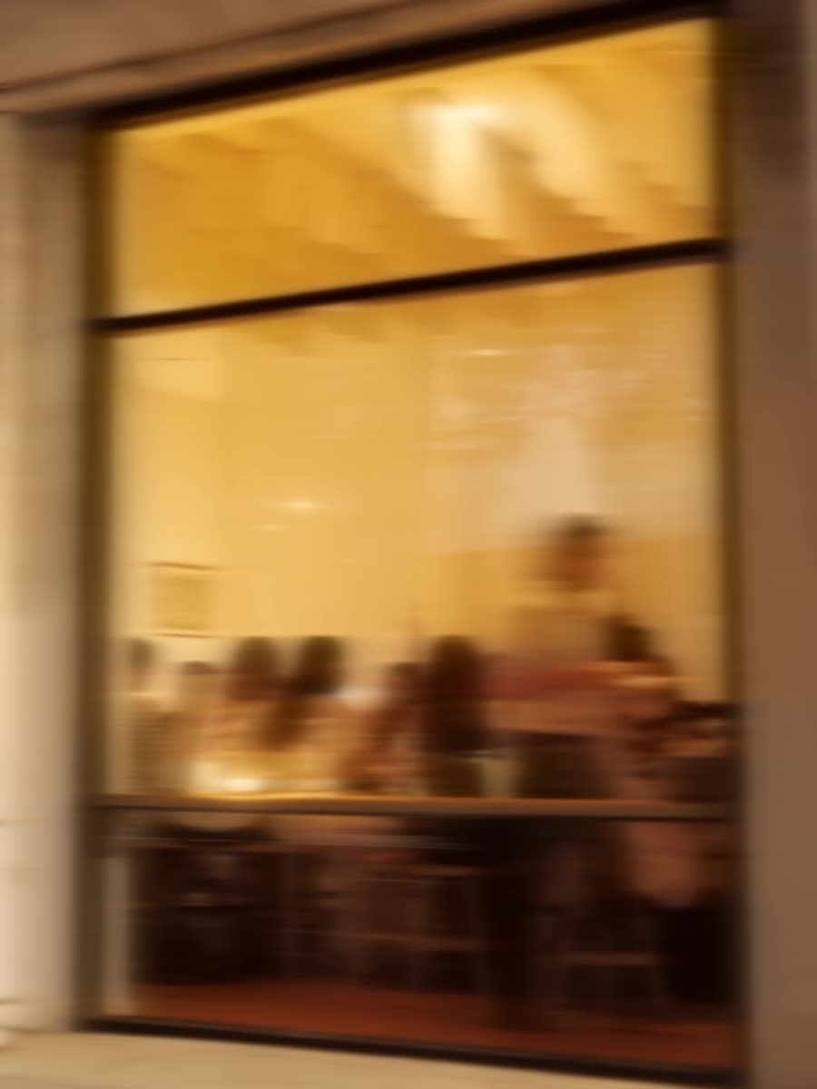 A long table of seated guests seen through a lit window at night, motion-blurred.
